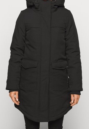 Parka d'hiver noire avec poches à rabat et à fermeture éclair, manches longues, capuche et poignets côtelés portée par une personne debout devant un fond neutre.