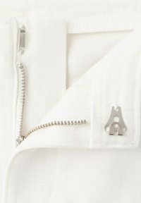Vue partielle d'un pantalon en denim blanc dézippé montrant une fermeture éclair en métal et un agrafe argenté à la taille.
