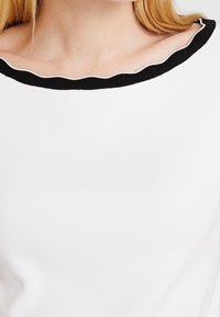Top blanco de canalé que presenta un escote negro con borde ondulado y detalles en blanco. La tela es suave y elástica, asegurando comodidad.