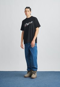 Black short-sleeve t-shirt with a metallic logo, paired with loose-fit blue jeans and beige boots. The background is a plain light color.