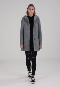 Cappotto grigio con zip e cappuccio, dalla texture morbida, maniche lunghe e vestibilità comoda, indossato sopra jeans neri strappati e sneakers nere.