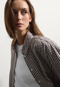 Camisa de cuadros marrón y blanca con cuello botón, fabricada en un tejido ligero y con una textura sutil, superpuesta a una camiseta blanca.