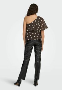 Femme aux cheveux ondulés portant un haut asymétrique noir à pois, associé à un pantalon en cuir noir et des bottines à talons noires, vue de dos.