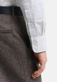 Camisa blanca con patrón azul de puntos, detalle de puños enrollados y botones dorados, combinada con pantalones marrones texturizados y un cinturón negro.