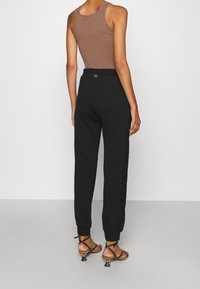 Žena ve černých teplácích se zlatým páskem, hnědém bezrukávém topu a černých sandálech na podpatku s pásky, stojící před jednoduchým pozadím.