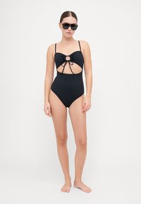 Maillot de bain une pièce noir avec design découpé, détail de nœud central, fines bretelles et jambes à haute coupe ; texture lisse et style minimaliste.