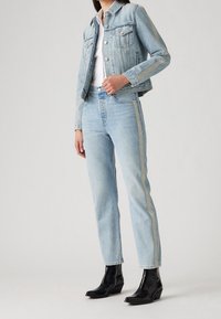 Světle modrá džínová bunda se dvěma předními kapsami a stříbrným pruhovaným detailem, v kombinaci s vysokými světle modrými džíny a černými kotníkovými botami.