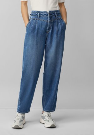 Frau trägt hoch taillierte, lockere blaue Jeans mit Falten, Vordertaschen und weißen Sneakers, stehend vor einem schlichten grauen Hintergrund.