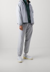 Chaqueta ligera gris claro, con capucha y cordón, combinada con joggers ajustados a juego. Zapatillas blancas con acentos de color completan el look.