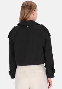 Donna con capelli castano chiaro lisci, che indossa una giacca nera corta con mostrine e pantaloni beige a vita alta, vista di schiena su sfondo bianco.