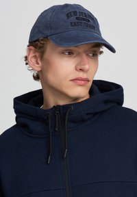 Felpa blu navale con zipper e cordini, caratterizzata da un tessuto testurizzato. Il cappellino coordinato presenta dettagli di testo cuciti.