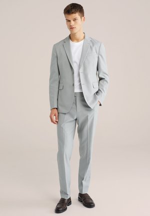 Bărbat tânăr purtând un costum gri deschis cu un tricou alb și mocasini maro închis, stând cu o mână în buzunar.