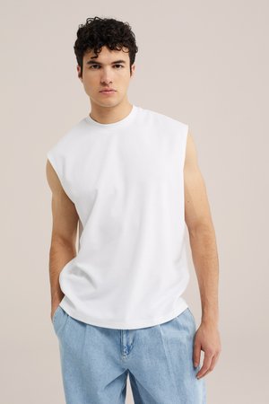Jeune homme aux cheveux bouclés portant un débardeur blanc et un jean bleu clair, debout devant un fond beige uni.