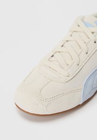 Sneaker in pelle scamosciata color crema con accenti blu chiaro e suola in gomma. Presenta lacci tradizionali e un logo vicino alla punta. Superficie testurizzata.