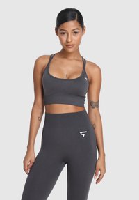 Mladá žena v tmavošedej športovej podprsenke a vysoko pásových legínach, stojaca s uvoľnenými rukami na svetlom pozadí.