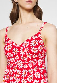 Czerwona sukienka kwiatowa z dekoltem w kształcie litery V, cienkimi ramiączkami typu spaghetti oraz wzorem biały stokrotek z żółtymi środkami. Miękki, lekki materiał.