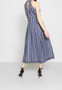 Marinblå klänning med rutigt mönster, ärmlös design och nyckelhålsdetalj i ryggen. Golvlång med utsvängd silhuett, kombinerad med svarta klackar.