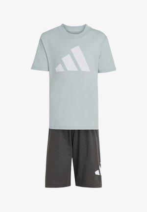 Due bambini indossano t-shirt grigie Adidas abbinate, pantaloncini neri, calzini bianchi Adidas e scarpe da ginnastica, in piedi davanti a uno sfondo semplice.