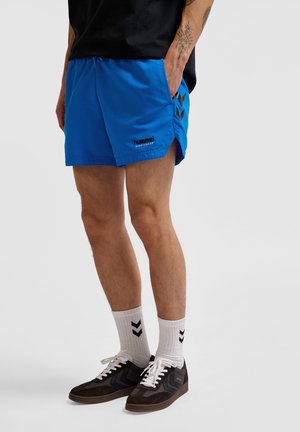 Blauwe sportshorts met zijsplitten, zwart logo, gedragen met witte sokken met een chevronpatroon en bruine sneakers met zwarte accenten.