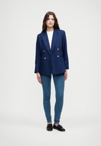 Mornarsko dvojno zapenjanje blazer z zlatimi gumbi, zareznimi lapelami, vertikalna silhueta, v kombinaciji s prilegajočimi se modrimi skinny jeans in črnimi čevlji.