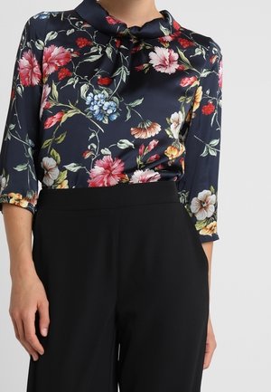 Woman wearing a navy floral blouse with red, pink, blue, and yellow flowers paired with high-waisted black pants.