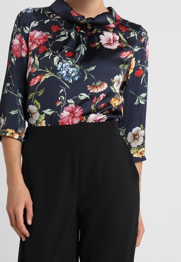 Femme portant un chemisier bleu marine à motifs floraux rouges, roses, bleus et jaunes, associé à un pantalon noir taille haute.