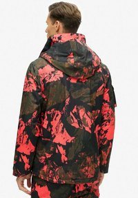 Chaqueta de camuflaje con capucha, con una base negra cubierta de patrones abstractos en rojo y marrón. Tejido texturizado, con mangas largas y bolsillos.