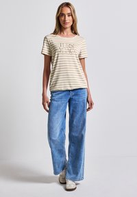 T-shirt rayé beige et blanc à manches courtes et col rond, associé à un jean large bleu clair. Design simple avec un texte imprimé.