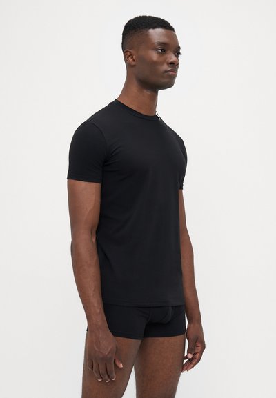 Schwarzes kurzärmliges T-Shirt aus weichem Stoff mit Rundhalsausschnitt. Getragen mit schwarzer enganliegender Unterwäsche steht das Modell im Profil vor einem schlichten Hintergrund.