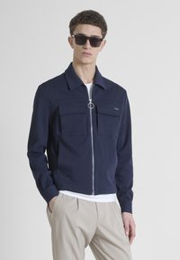 Giacca zip-up navy con due tasche sul petto, colletto piatto e zip con anello in metallo. Indossata sopra una camicia bianca e pantaloni beige.