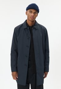 Vyras, vilkintis tamsiai mėlyną, sagotuotas paltą ir megztą kepurę, stovintis priešais paprastą baltą foną.