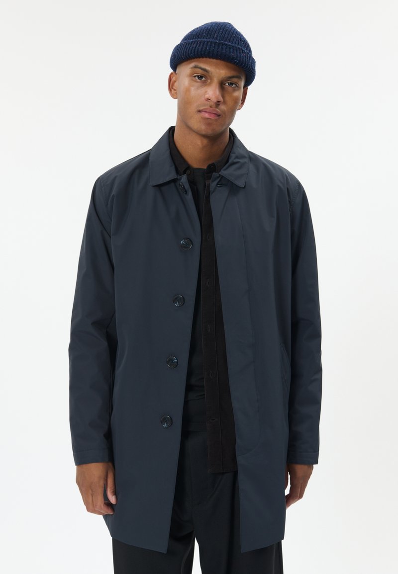 Vyras, vilkintis tamsiai mėlyną, sagotuotas paltą ir megztą kepurę, stovintis priešais paprastą baltą foną.