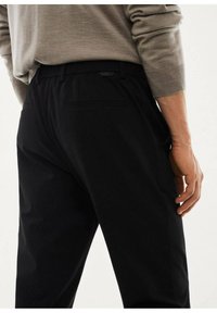 Pantaloni neri con una texture liscia, dotati di una cintura e tasche posteriori. Un'etichetta con il logo è visibile sul retro.