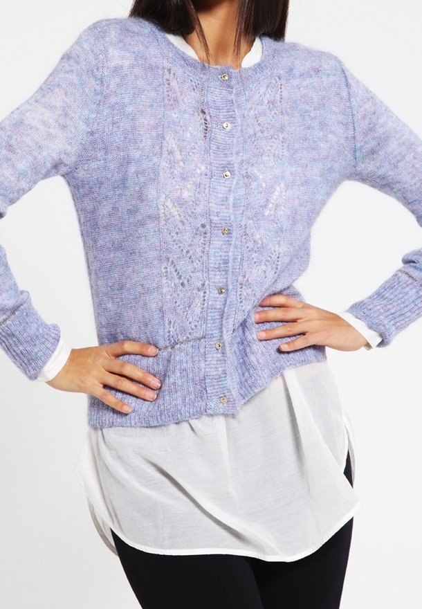 Femme portant un cardigan en tricot lavande boutonné sur une longue chemise blanche et un pantalon noir, les mains sur les hanches, posant devant un fond uni.