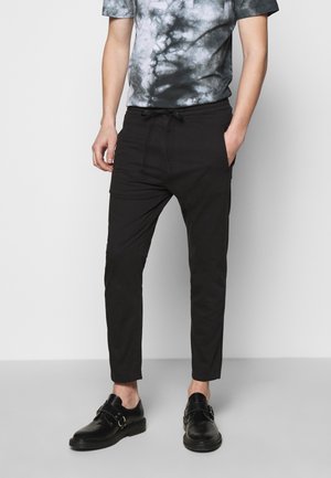 Pantalons noirs ajustés avec une taille élastique et des poches latérales, associés à un t-shirt gris tie-dye et des chaussures noires à boucles.