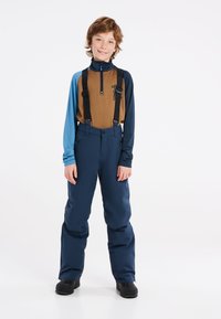 Ski-outfit met een bruin en blauwe lange mouwen top met rits en zwarte bretels, gecombineerd met marineblauwe skibroeken en zwarte laarzen.