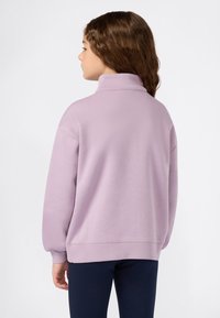 Jeune personne avec des cheveux bruns ondulés portant un sweat à col roulé lilas et un pantalon sombre, se tenant dos à un fond uni.
