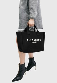 Fekete vászontáska, masszív fogantyúkkal; a "ALLSAINTS LONDON" fehér betűkkel van nyomtatva. Fekete bokacsizmával viselve.