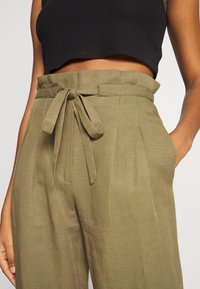 Pantalon taille haute vert olive avec ceinture nouée, devant plissé et poches latérales, confectionné dans un tissu texturé.