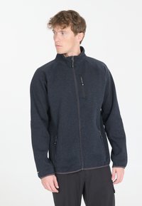 Tummansininen fleece-takki korkealla kauluksella, täysipitkällä vetoketjulla ja kahdella vetoketjullisella sivutaskulla. Takissa on sileä pinta ja joustavat hihansuut.