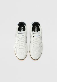 Sneakers bianchi con tomaia in pelle texture, punta perforata, dettagli neri e suola in gomma color caramello; presentano lacci bianchi piatti e dettagli del logo.