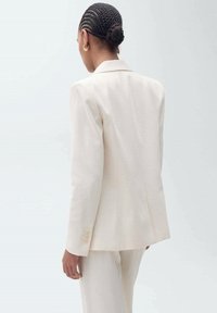 Blazer beige clair sur mesure avec un design structuré, fermeture à bouton unique et accents à trois boutons aux poignets. Tissu lisse et lignes nettes.