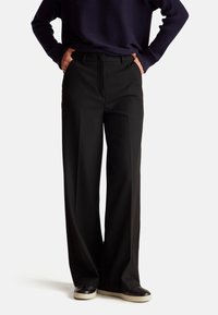 Wide-leg black trousers made of smooth fabric, featuring a front crease, belt loops, and side pockets, paired with dark sneakers.