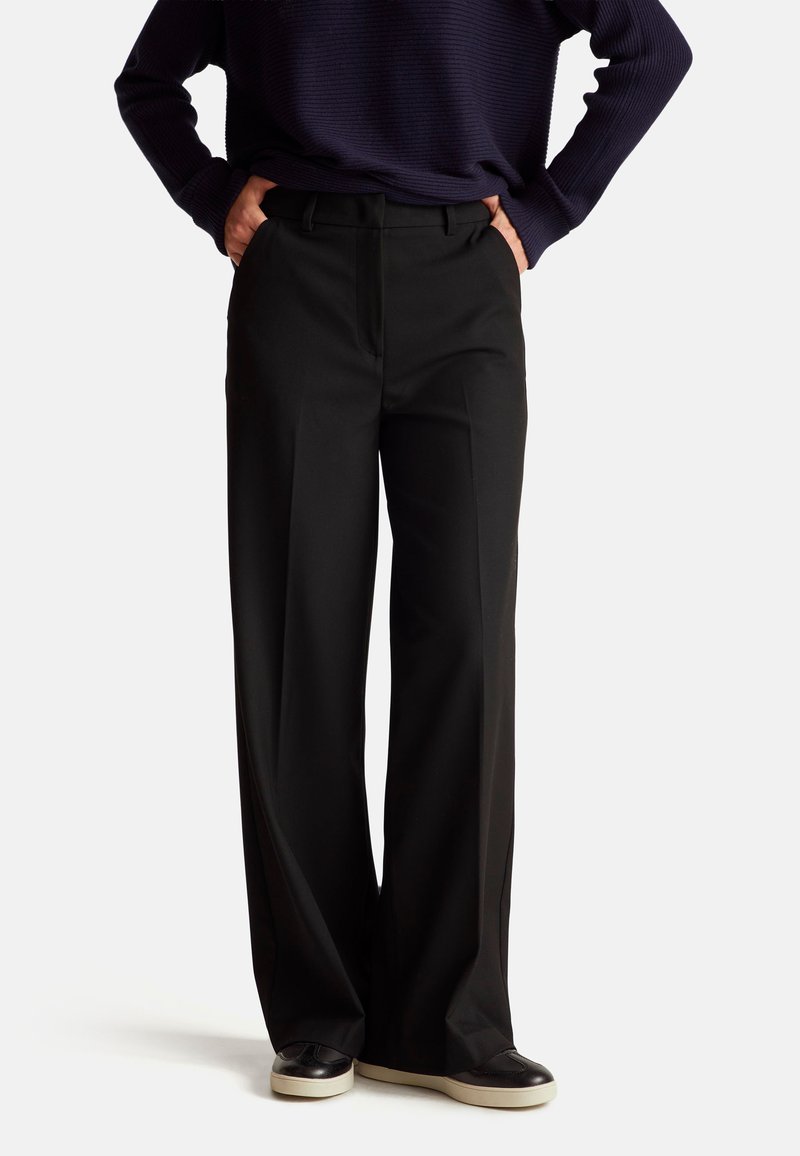 Wide-leg black trousers made of smooth fabric, featuring a front crease, belt loops, and side pockets, paired with dark sneakers.