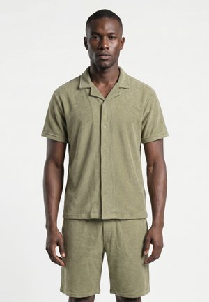 Homme portant une chemise boutonnée à manches courtes vert olive clair assortie et un short mi-cuisse, debout devant un fond blanc uni.