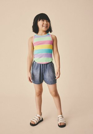 Fiatal lány mosolyog, szivárványszínű csíkos ujjatlan felsőt, farmer rövidnadrágot és fehér szandált visel, egyszerű bézs háttér előtt áll.