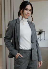 Szary żakiet w wzór houndstooth z klapami, dwoma guzikami i bocznymi kieszeniami, zestawiony z białym golfem oraz pasującymi szarymi spodniami.