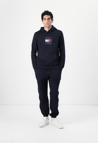 Sweat à capuche bleu marine avec une poche avant, un graphique de logo et un pantalon de survêtement assorti. Les deux ont une texture douce et des poignets côtelés. Des baskets blanches complètent le look.