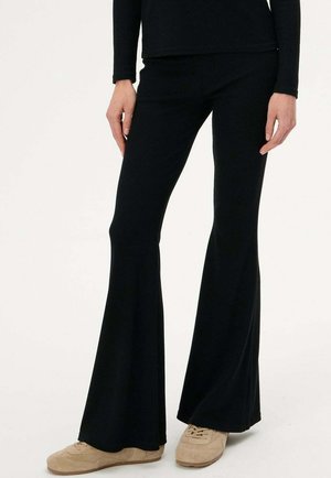 Osoba w czarnych dzwonowanych spodniach i beżowych zamszowych butach sportowych, stojąca na jasnym, jednolitym tle.