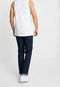 Débardeur blanc, jean bleu foncé et baskets blanches. Le jean est retroussé au niveau des ourlets, créant une coupe décontractée et un style relax.